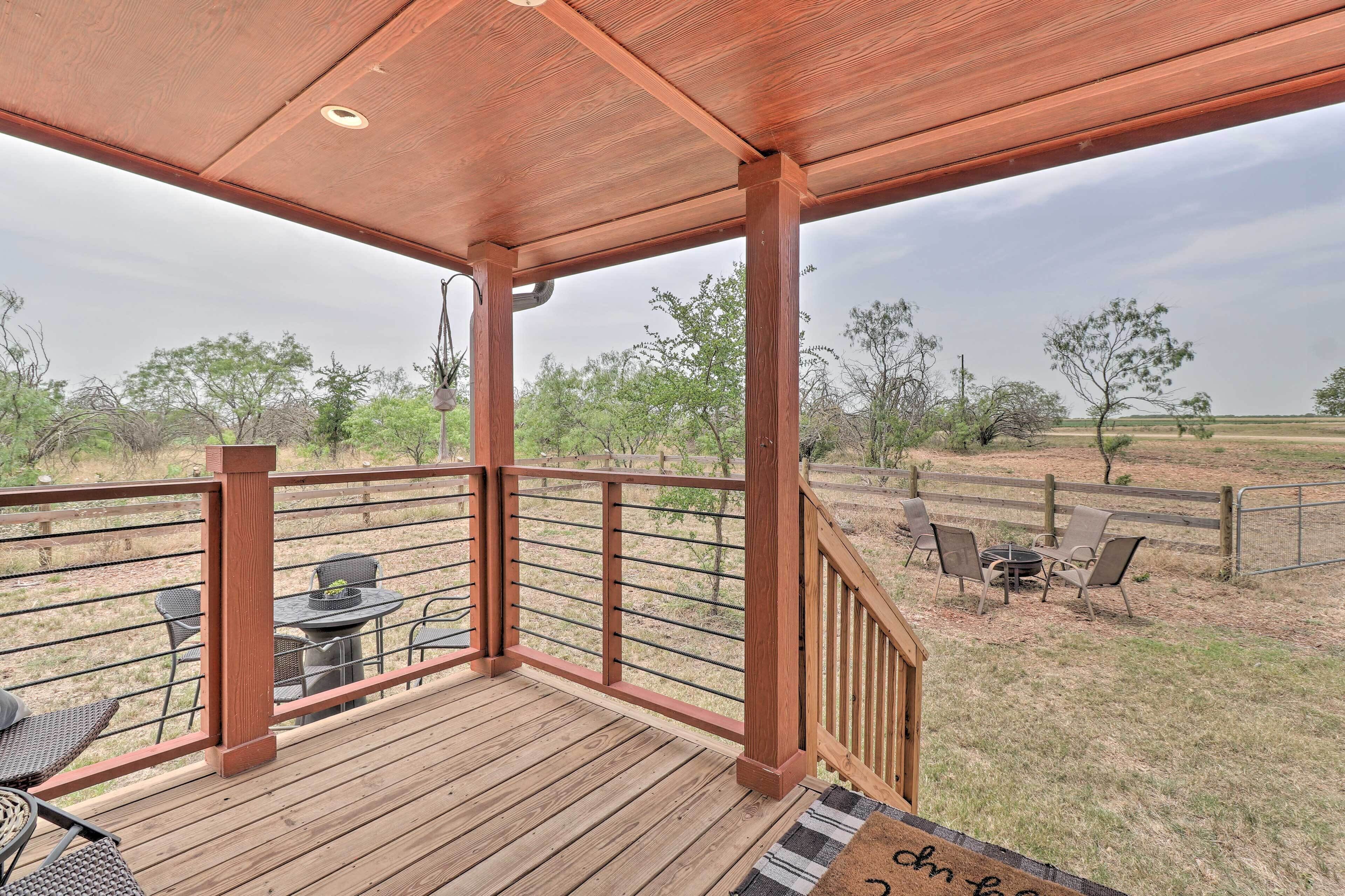 Une terrasse en bois avec une rambarde, donnant sur un espace clos aménagé avec des sièges d’extérieur et des arbres.