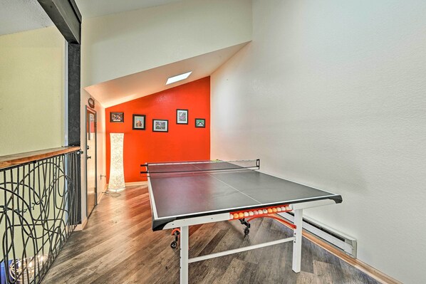 A ping pong table in a room with wooden flooring, a red accent wall, and a staircase with a metal railing. - Relaxing Deck & Grill: Pinetop Townhome! (Pinetop)