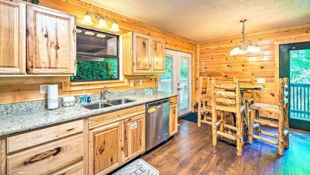 A wooden cabin-style kitchen with granite countertops, wooden cabinets, a stainless steel dishwasher, and a dining area with wooden chairs. - Cozy Gatlinburg Hideaway w/ Private Hot Tub! (Gatlinburg)