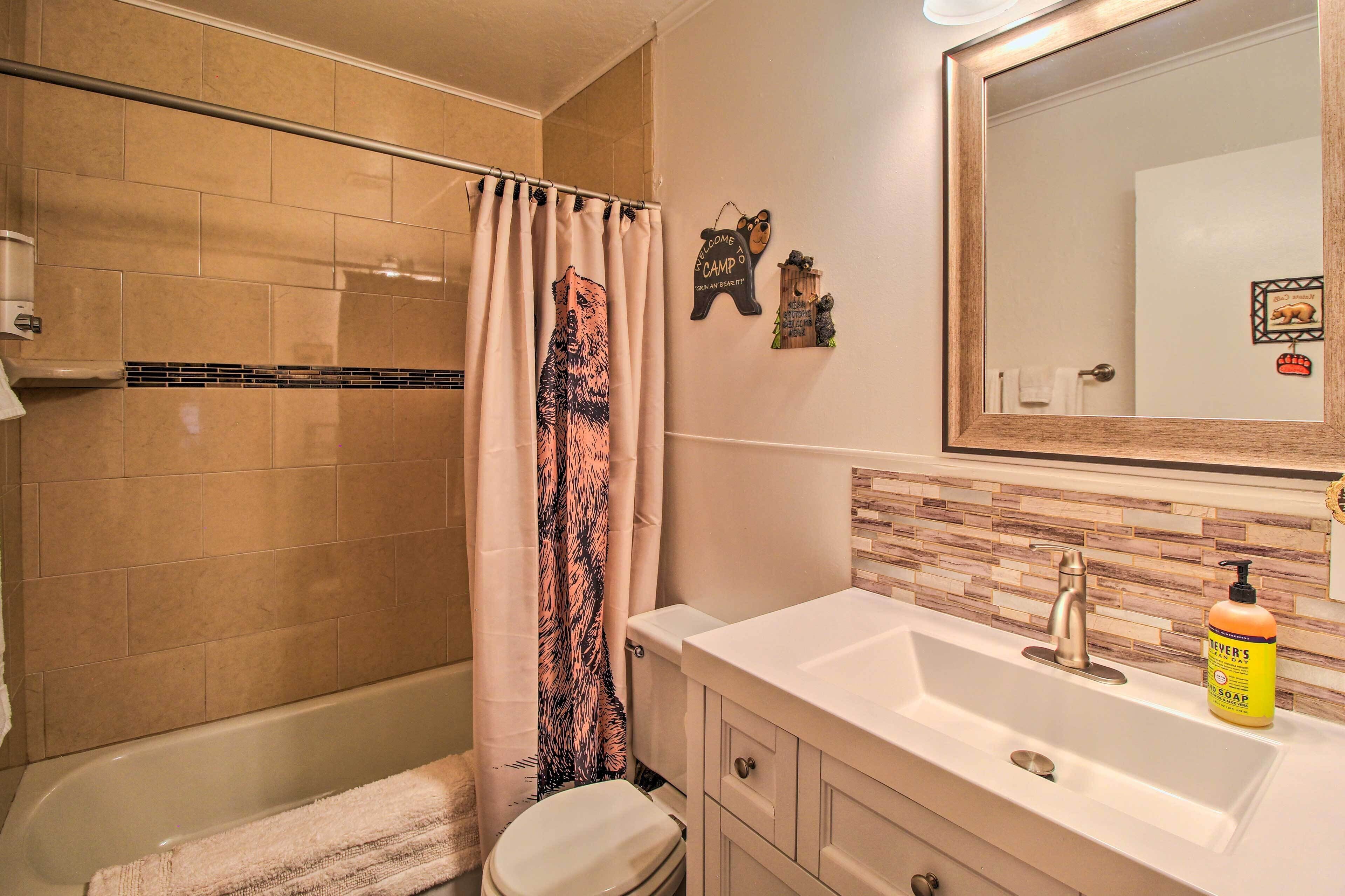 Un baño con baldosas color beige, una cortina de ducha con diseño de oso, un lavabo con dispensador de jabón y un espejo.