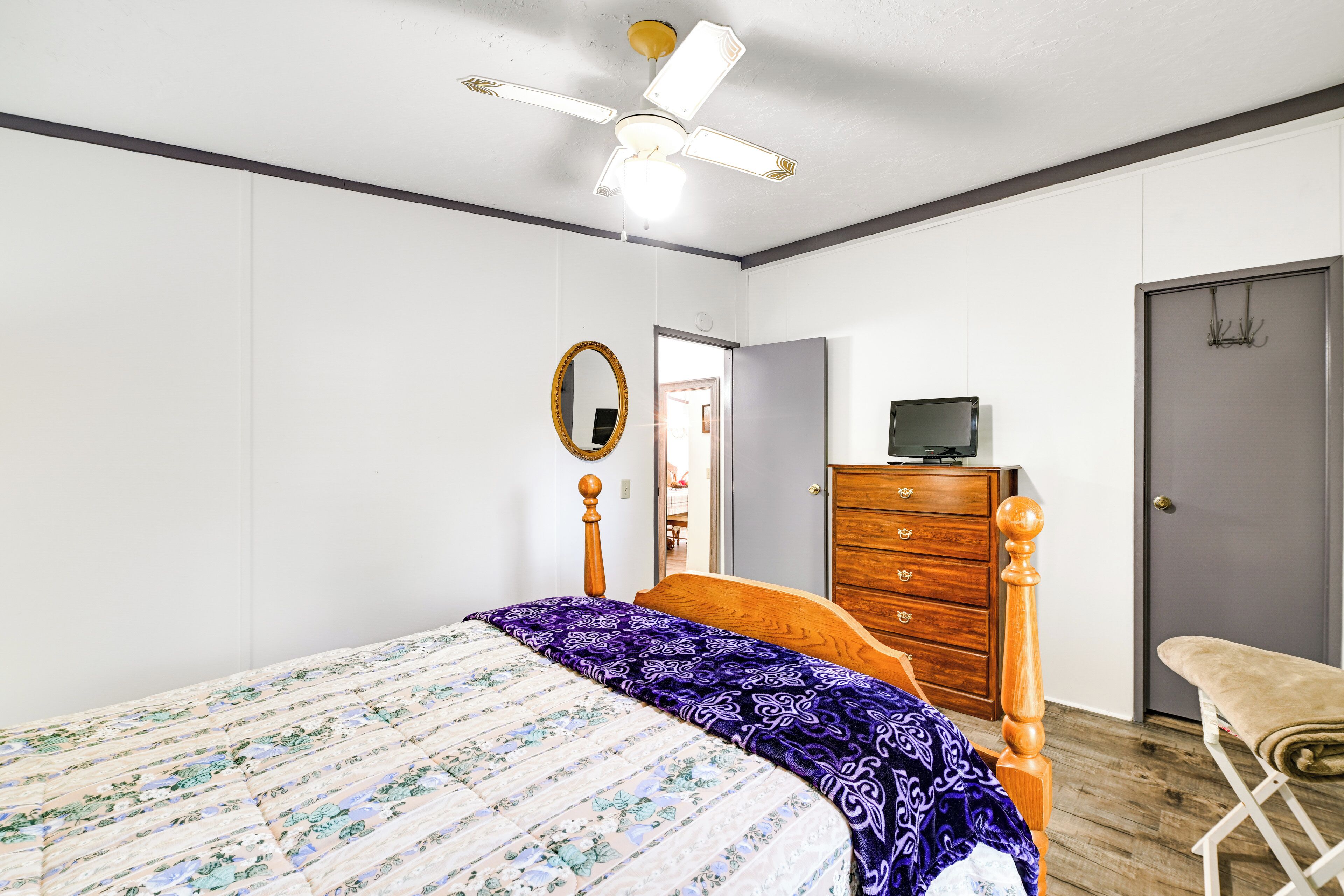 Une chambre à coucher comprenant un lit, une commode en bois, un miroir, un ventilateur de plafond et une porte.