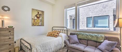 Une chambre à coucher comprenant un lit, un canapé, une commode, une fenêtre avec des stores et une œuvre d’art accrochée au mur.
