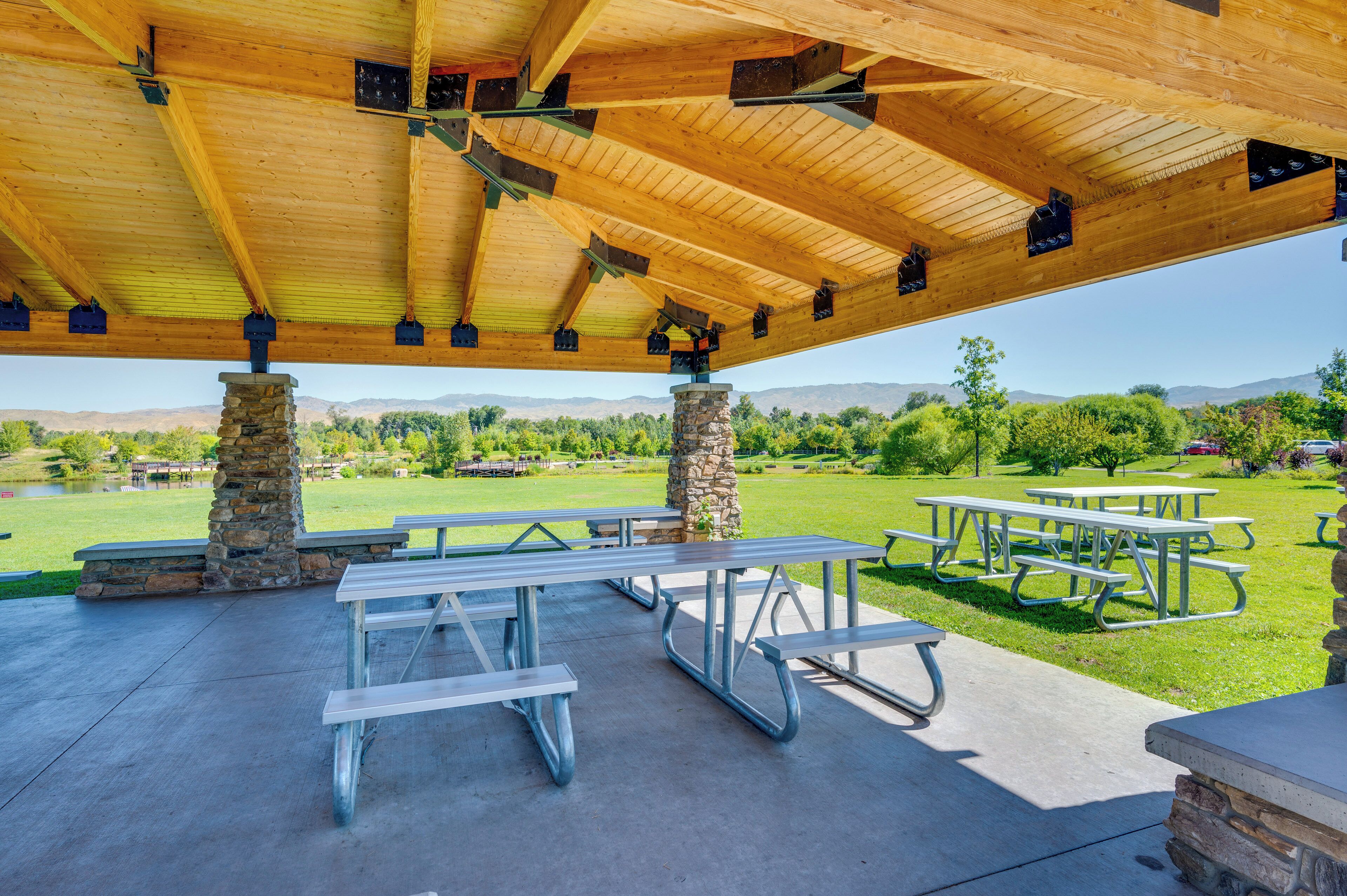 An outdoor pavilion with picnic tables and stone pillars, set against a backdrop of green fields and distant mountains.
