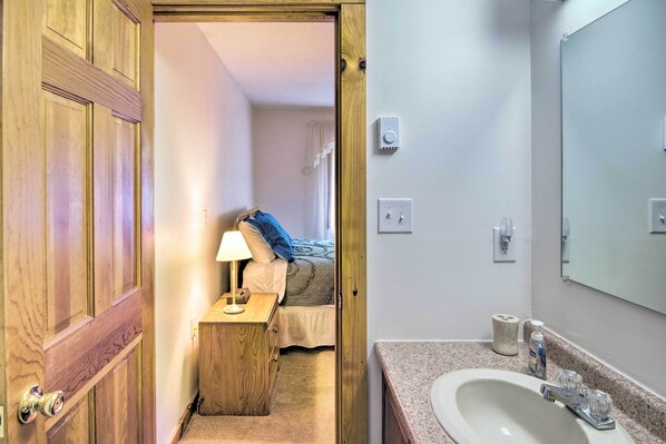 Una habitación de hotel con una puerta de madera, una mesita de noche con una lámpara, una cama con un almohón azul y un baño con lavamanos y espejo.
