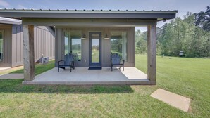 A covered porch with two Adirondack chairs, a concrete floor, and a view of a grassy yard and trees.