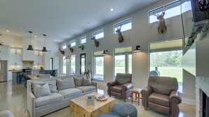 A spacious living room with a large sofa, two leather armchairs, and a coffee table. There are multiple windows providing natural light and views of the outdoors.