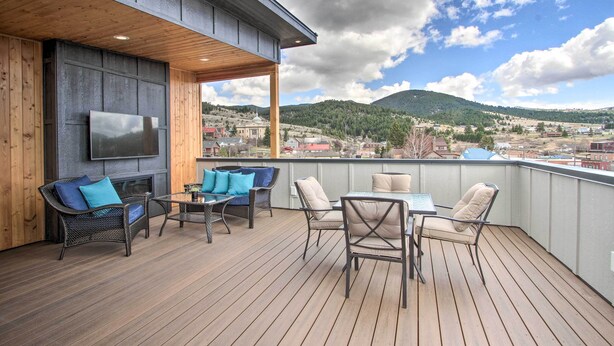 A wooden deck with outdoor furniture, a flat-screen TV, and a view of a town and mountains. - Exquisite Discovery Mtn Home w/ Sweeping Views! (Philipsburg)