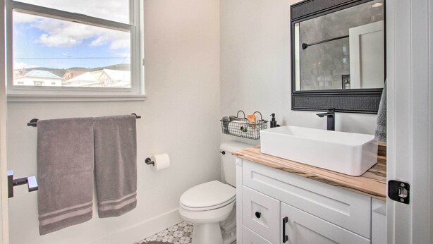 A bathroom with a toilet, a sink with a mirror above it, and a window with a view of houses. - Exquisite Discovery Mtn Home w/ Sweeping Views! (Philipsburg)