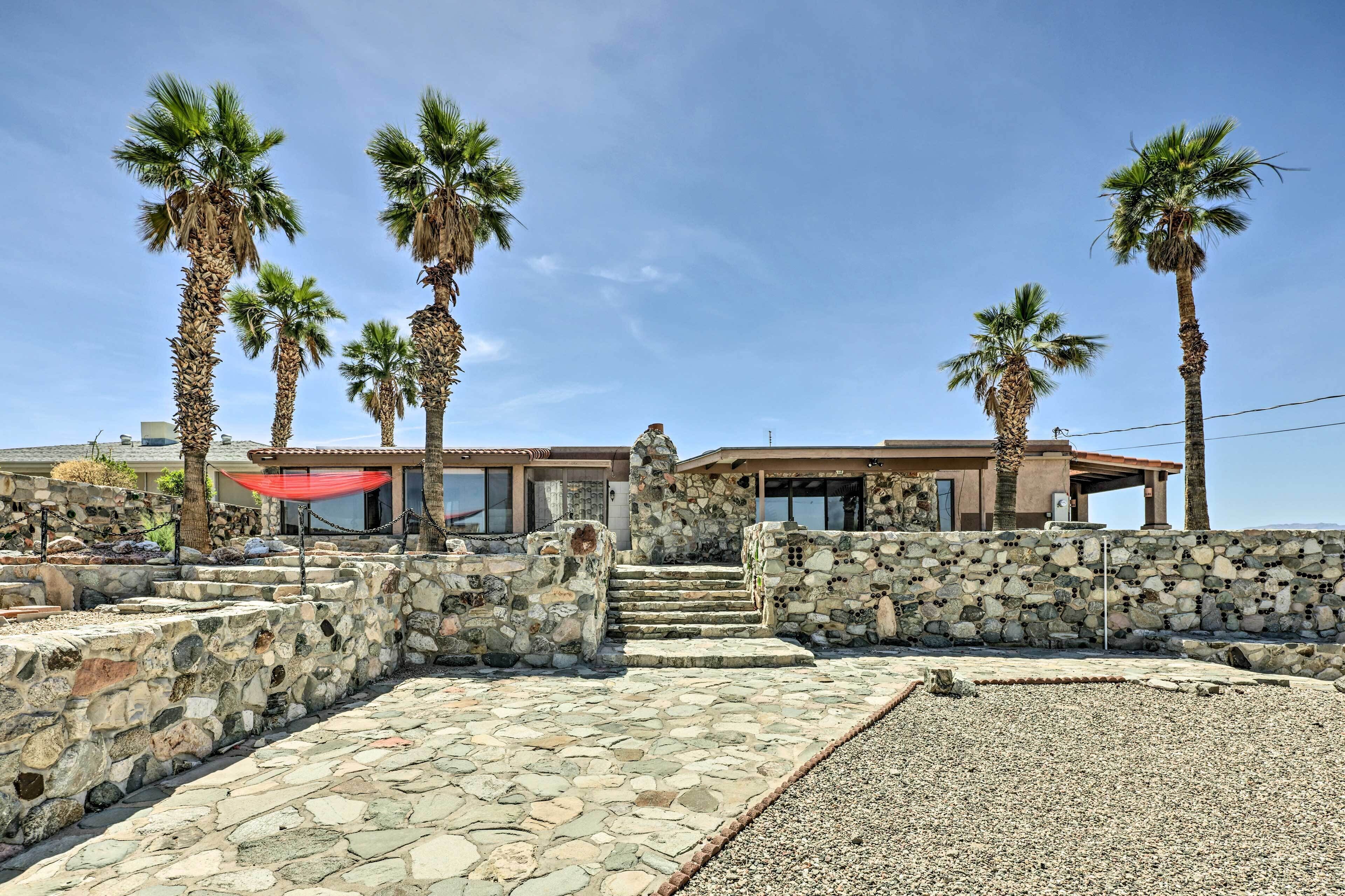 A stone-walled building with a stone staircase leading to a door, surrounded by palm trees.