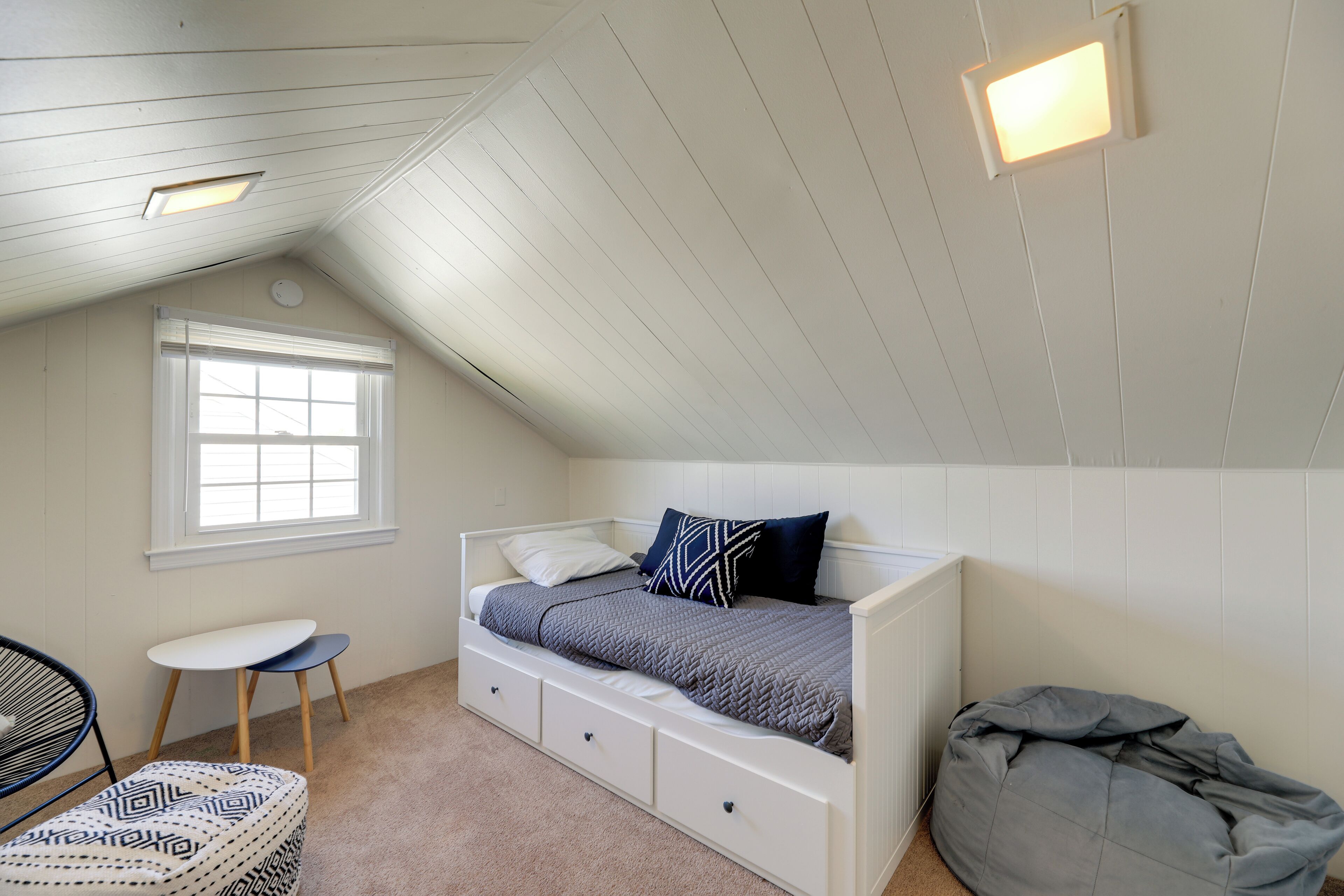 A bedroom with a sloped ceiling, a single bed with storage drawers, a window, a chair, and a bean bag.