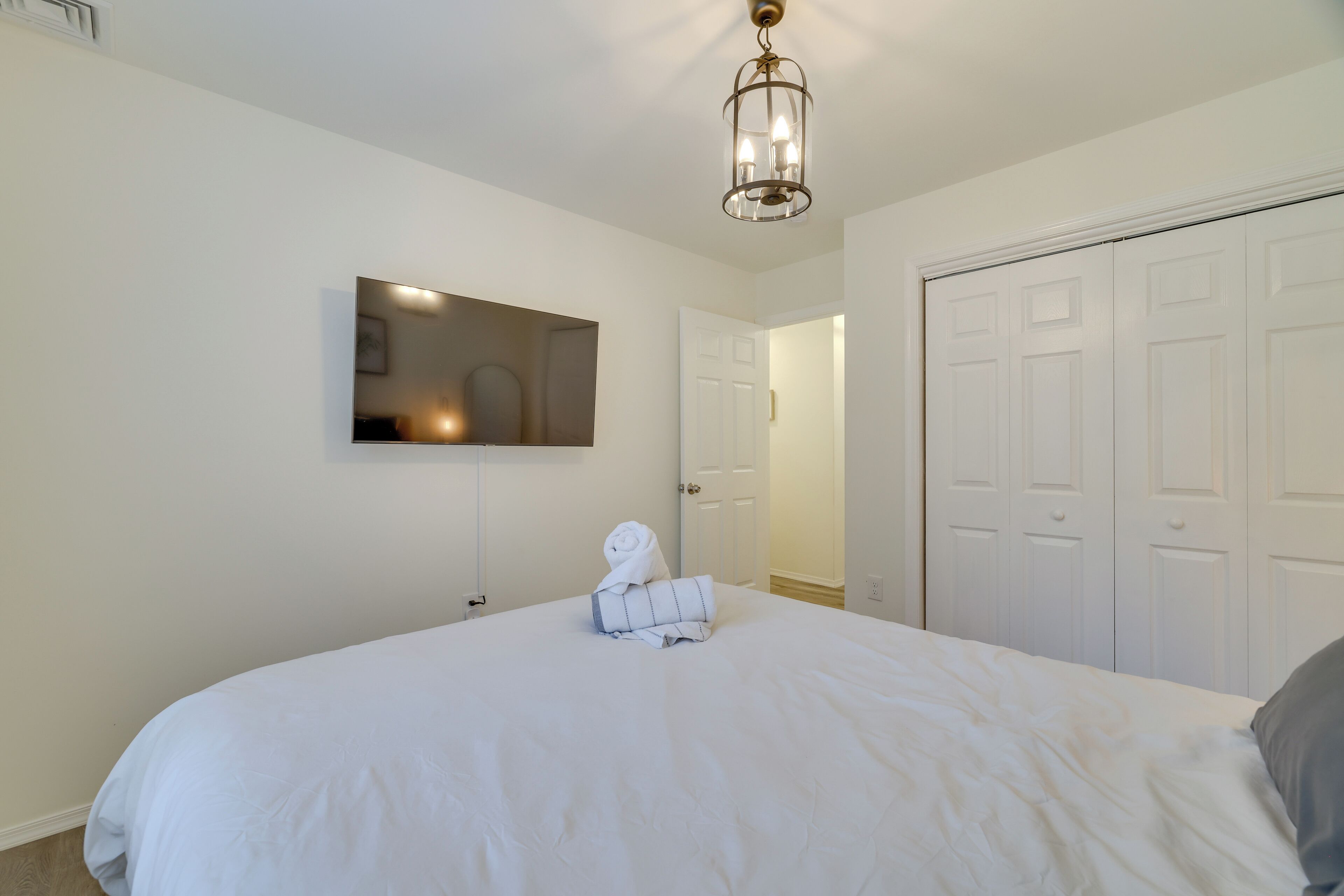 A neatly made bed with a white comforter and a rolled towel, a mounted flat-screen TV, and a white door with a handle.