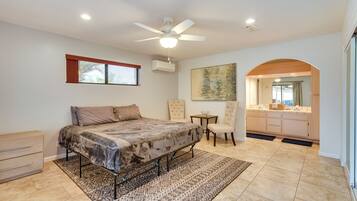A bedroom with a bed, a ceiling fan, a window with curtains, an air conditioning unit, a small table with a chair, and a view into the bathroom.