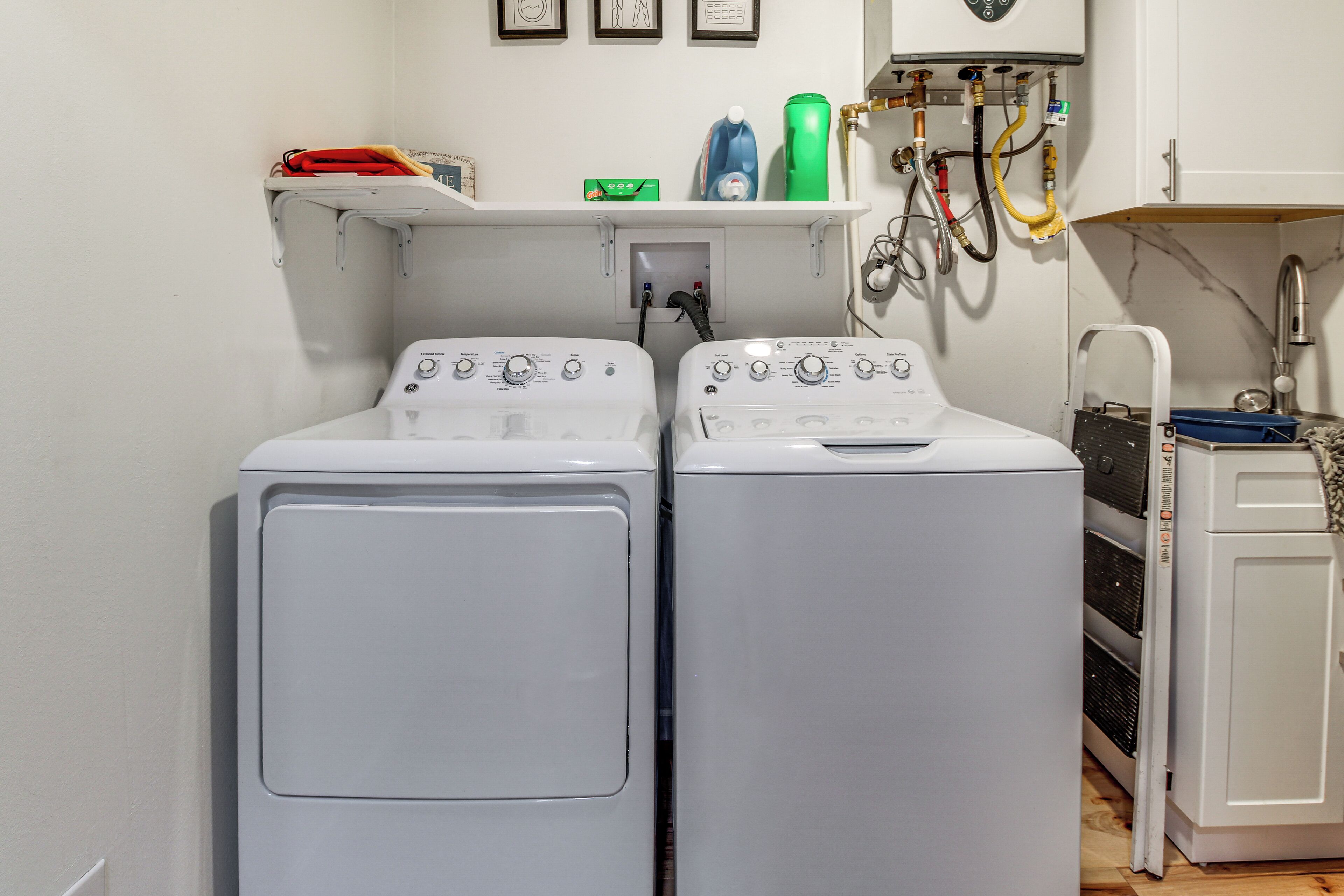 A laundry room with two front-loading washing machines, a shelf with cleaning supplies, and a water heater.