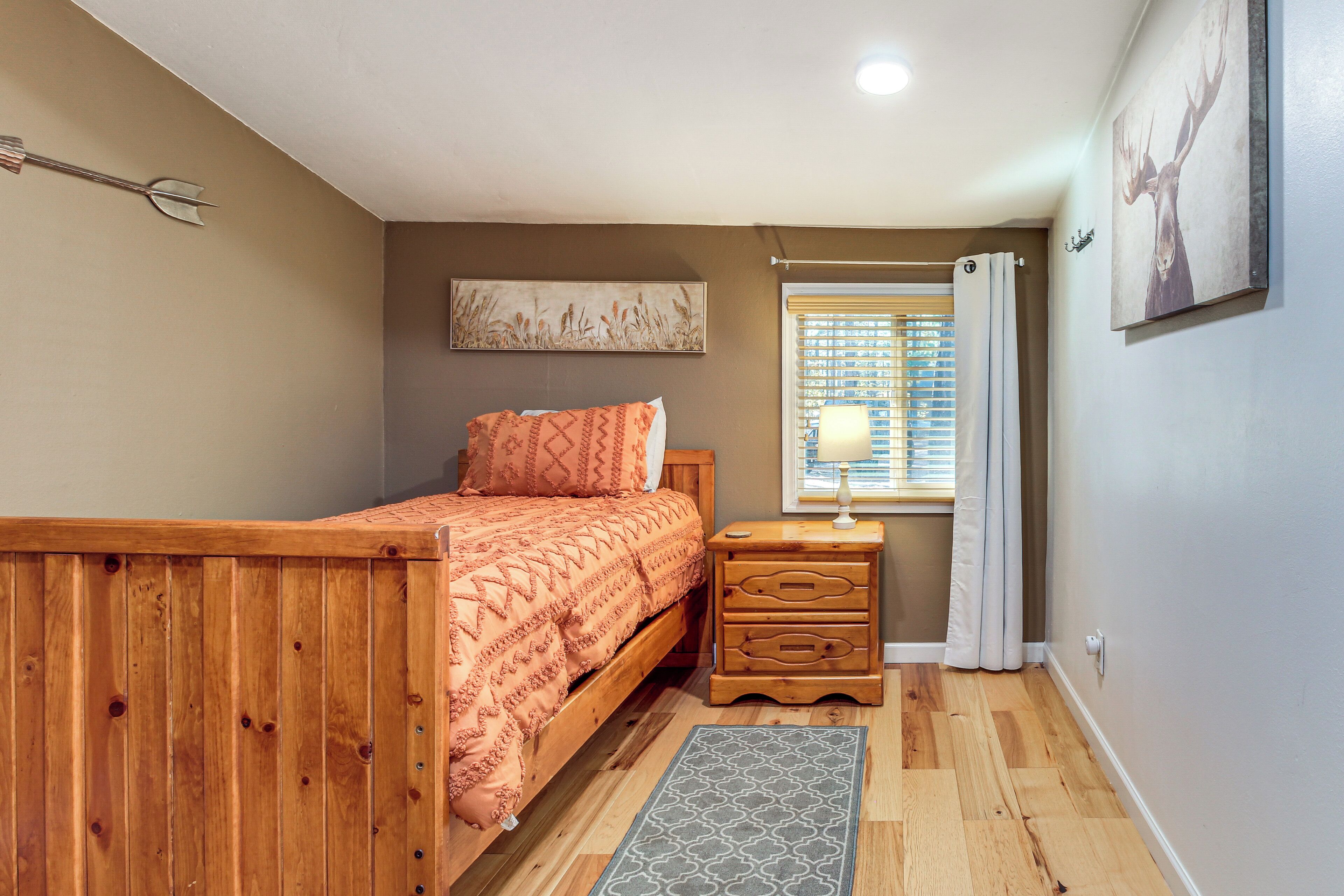 A bedroom with a wooden bed, a nightstand with drawers, a window with blinds, and a wall-mounted artwork.