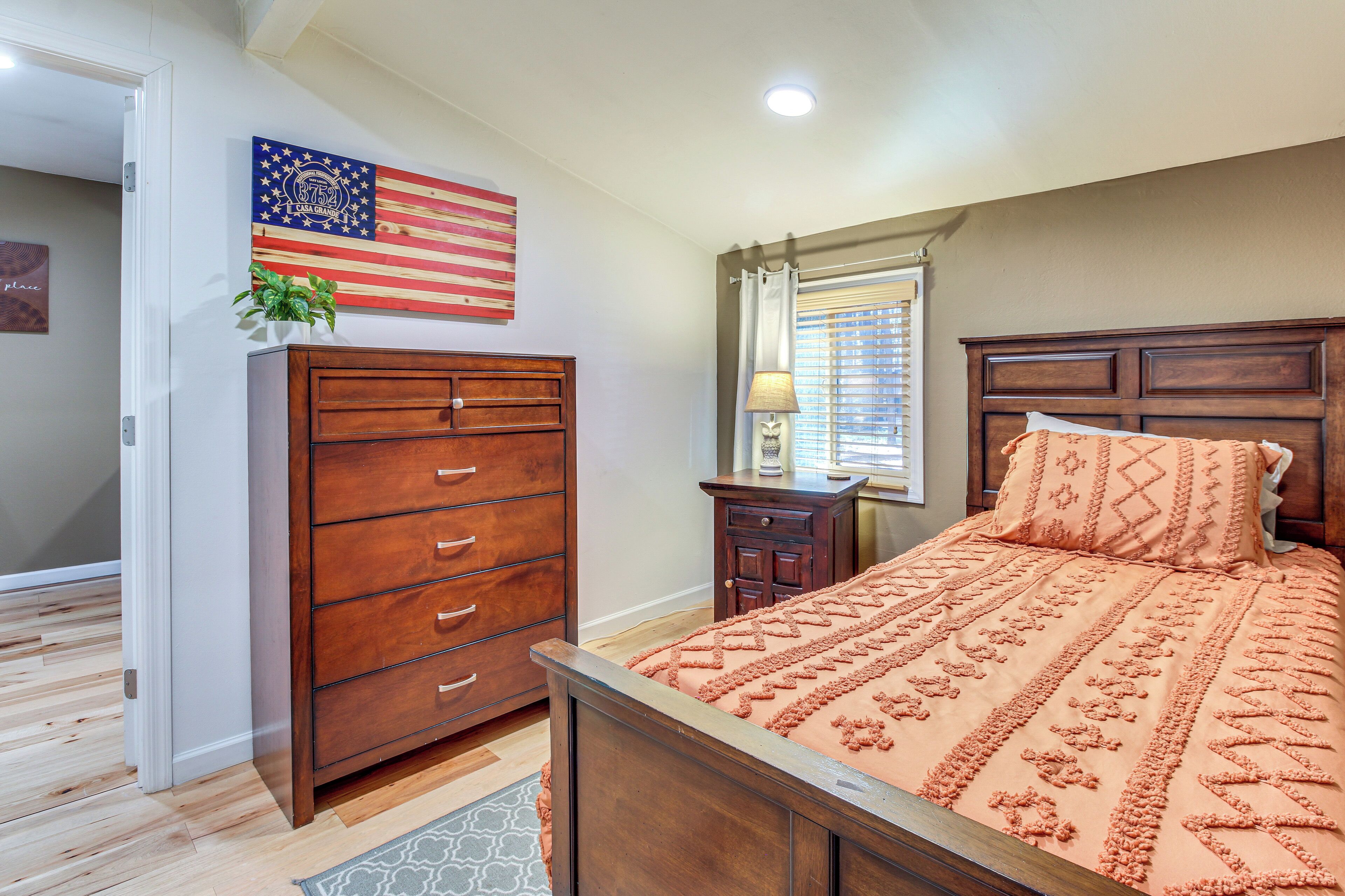 A bedroom with a wooden bed, a dresser, a nightstand, an American flag decoration, and a window with curtains.