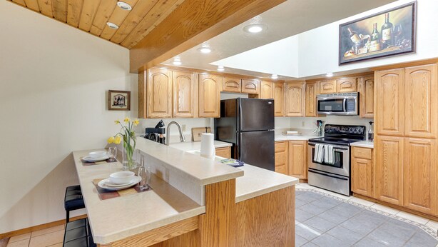 A modern kitchen with wooden cabinets, a central island, stainless steel appliances, and a dining area with chairs. - Nampa Vacation Rental Townhome Near Lake Lowell! (Nampa)