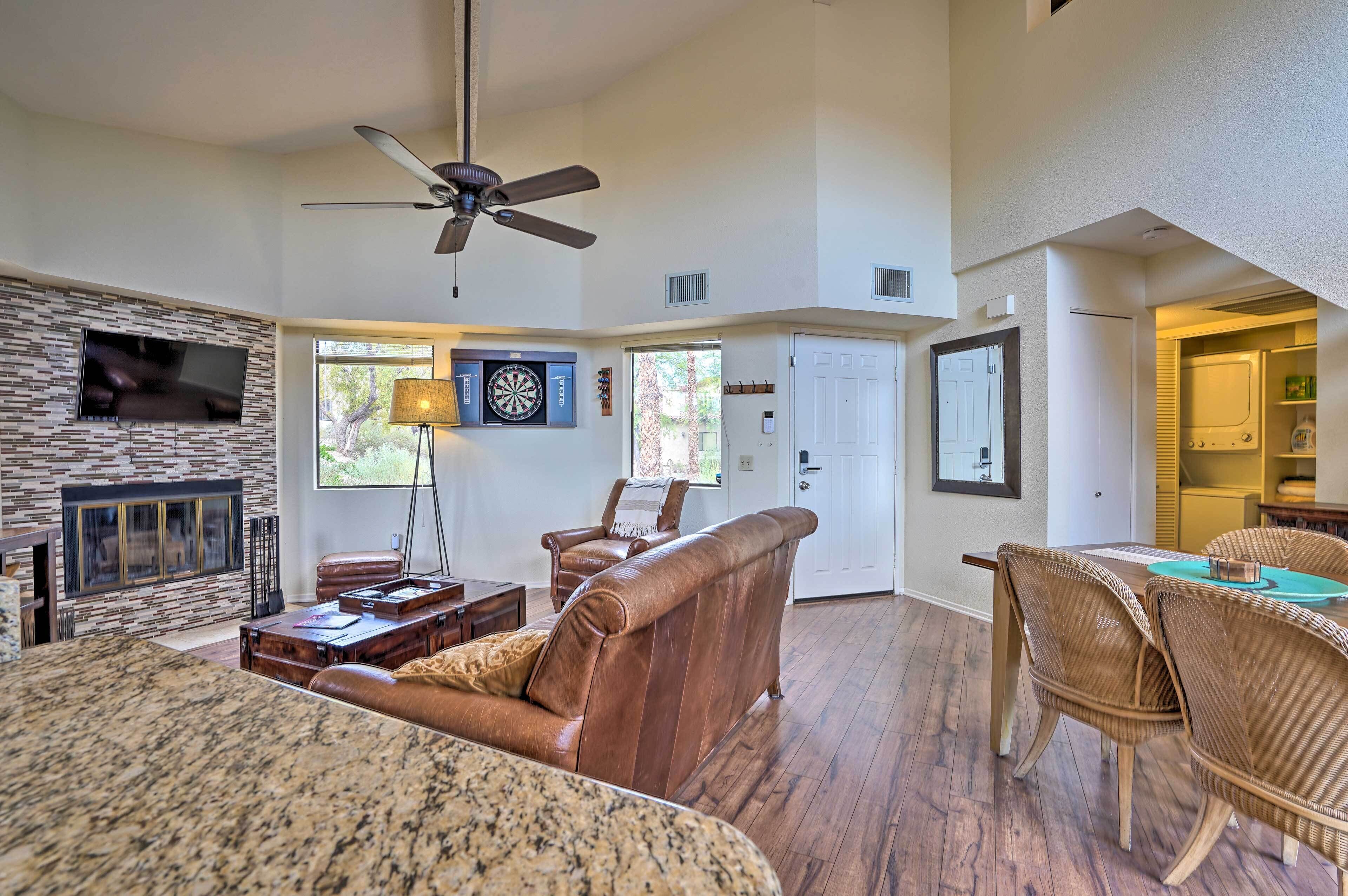 A spacious living room with a fireplace, a ceiling fan, a dartboard, and a dining area with wicker chairs.