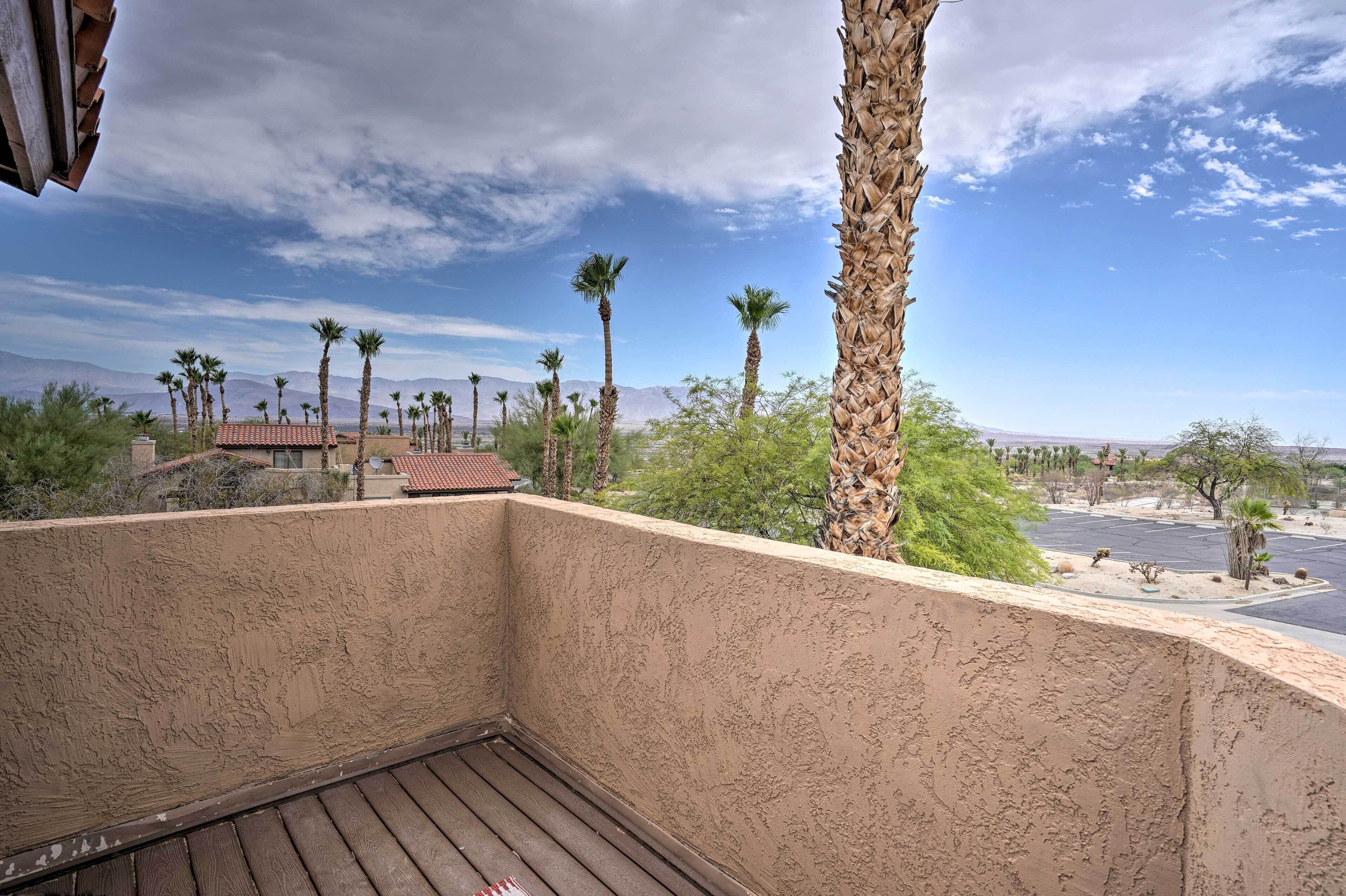 A balcony with a view of palm trees, a parking lot, and distant mountains under a partly cloudy sky.