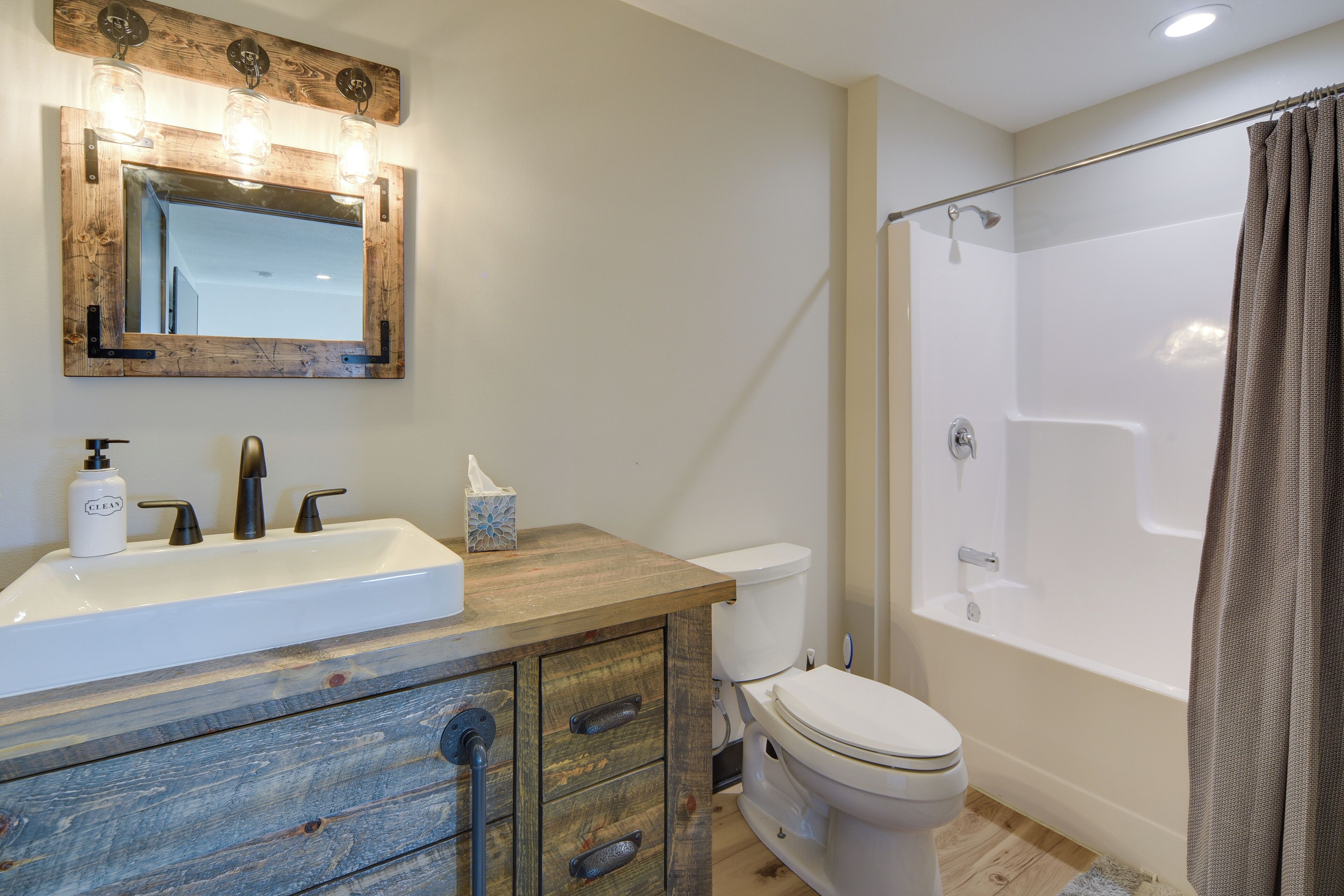 A bathroom with a wooden vanity, a white sink, a mirror with lights, a toilet, and a shower with a curtain.