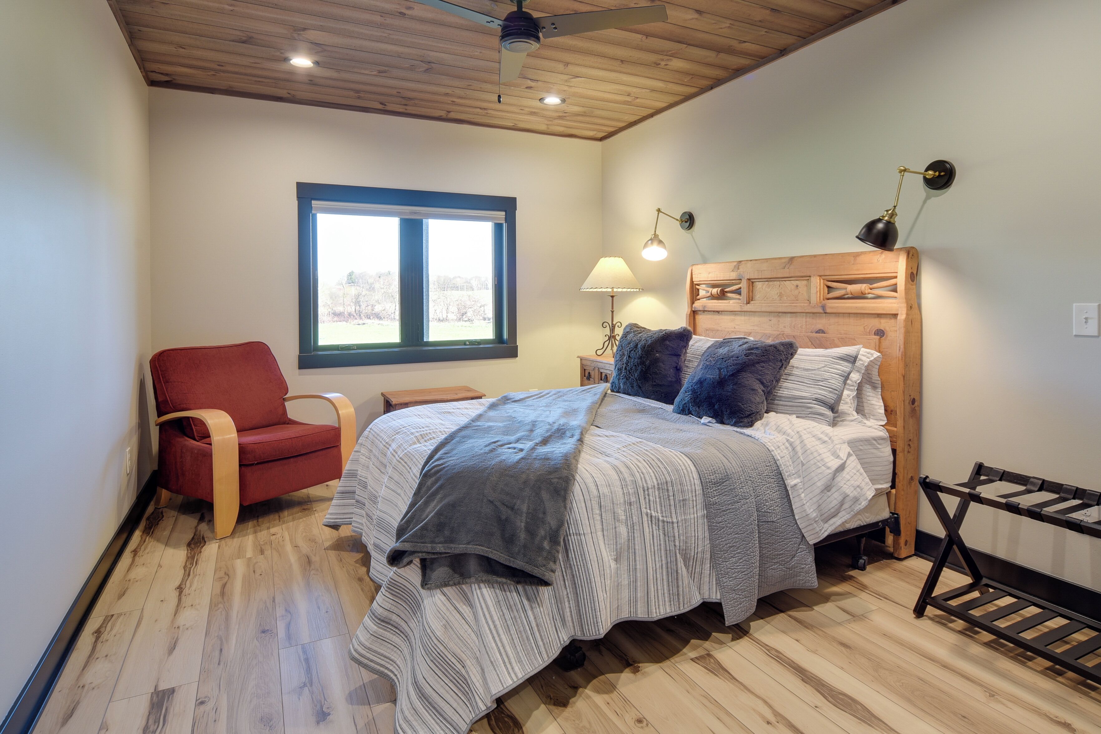 A bedroom with a wooden ceiling, a bed with a striped bedspread, a red chair, a window with a view, and a lamp on the nightstand.