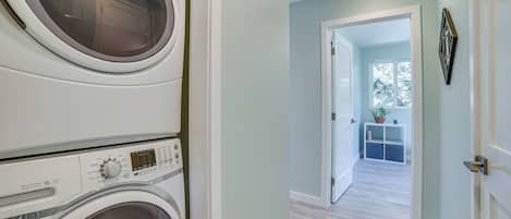 Une buanderie avec des lave-linge et des sèche-linge superposés, un couloir menant à une pièce dotée d’une fenêtre et d’une plante.