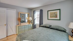 Une chambre à coucher avec un lit, une commode, un miroir, une fenêtre avec des rideaux et un tableau encadré au mur.