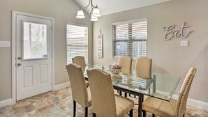 A dining area with a glass table surrounded by four upholstered chairs, a chandelier above, and a wall sign that reads "Eat".