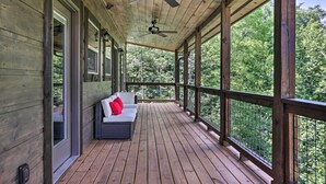 A wooden deck with a ceiling fan, a sofa with a red pillow, and a view of trees through large windows.