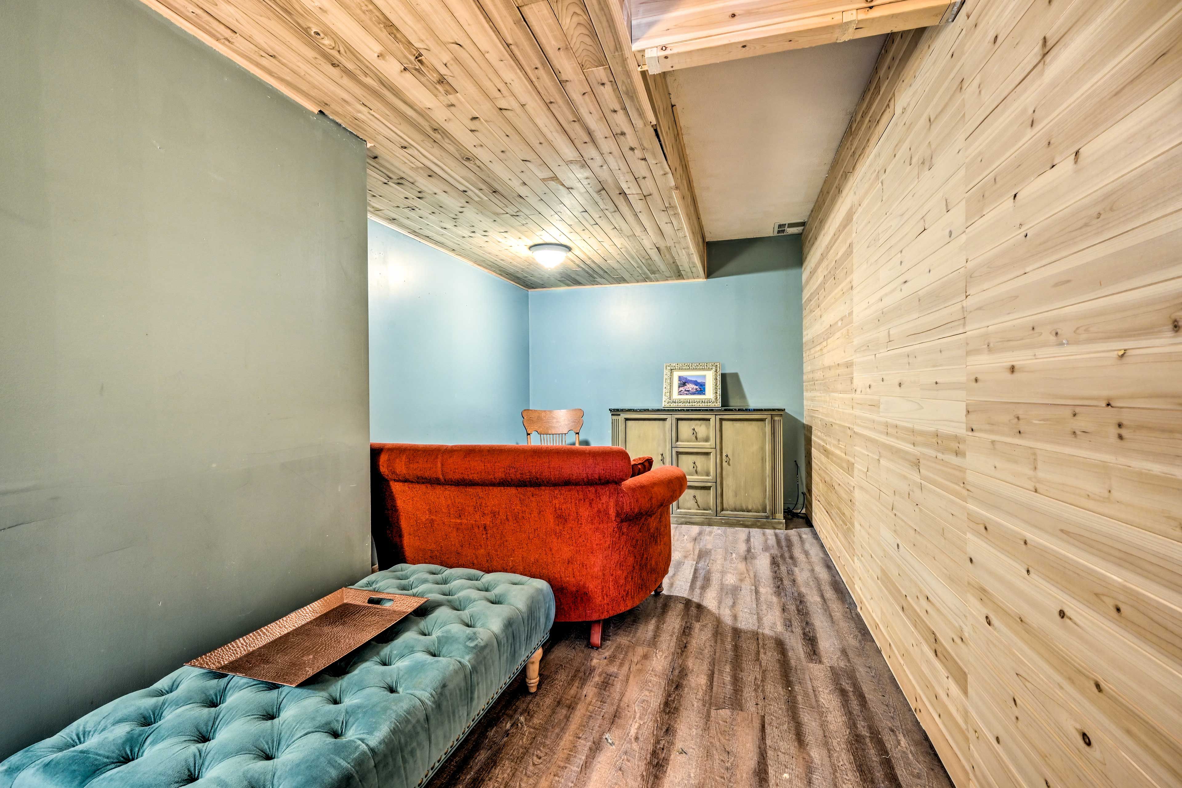A room with a wooden ceiling, a blue wall, a red armchair, a blue tufted ottoman, and a wooden cabinet.