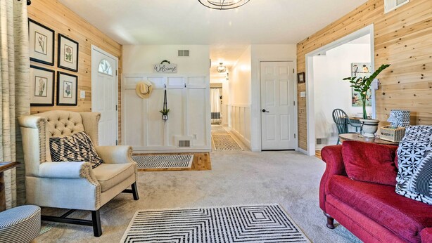 Un salon avec un fauteuil beige, un canapé rouge et un tapis à motifs. On y trouve des tableaux encadrés au mur, une pancarte « Bienvenue » et un lustre.