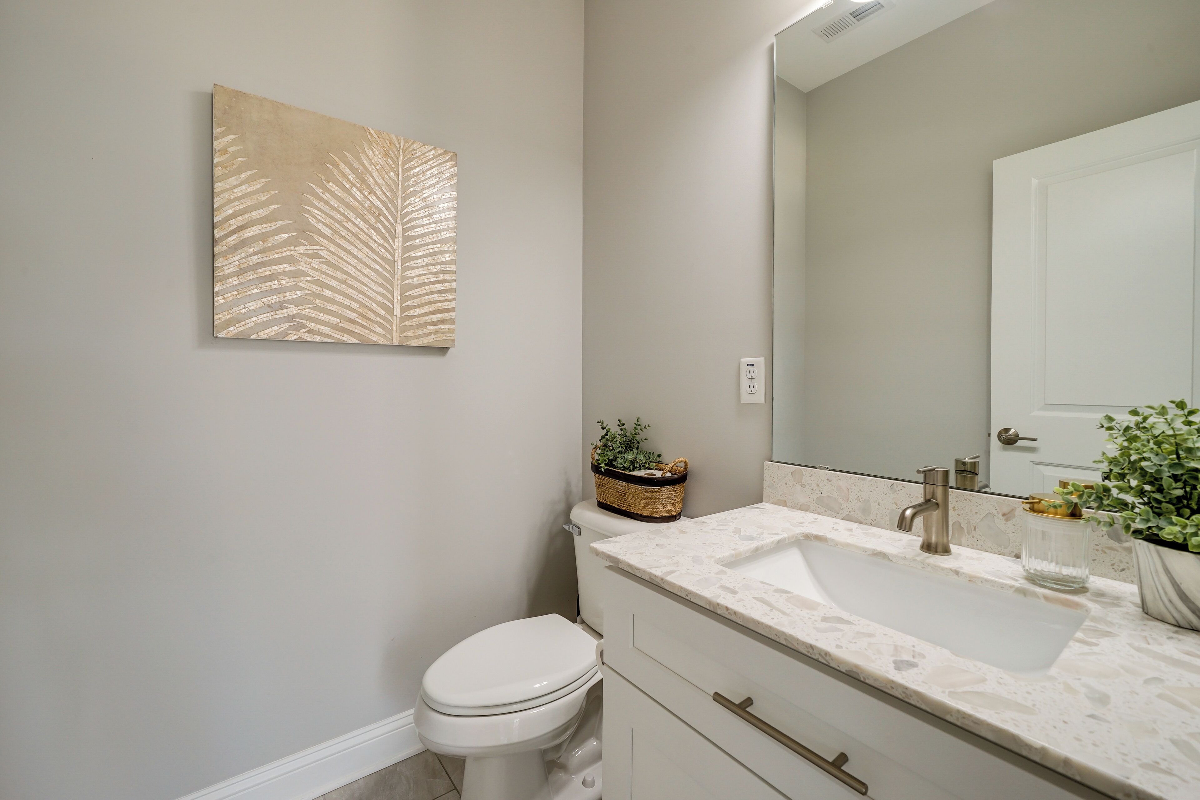 Une salle de bain avec des toilettes blanches, un lavabo doté d’un plan de travail en marbre, un grand miroir et une œuvre d’art représentant une feuille, accrochée au mur.