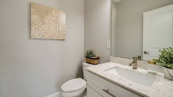 Une salle de bain avec des toilettes blanches, un lavabo doté d’un plan de travail en marbre, un grand miroir et une œuvre d’art représentant une feuille, accrochée au mur.