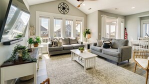 A spacious living room with a high ceiling, large windows, a ceiling fan, and a view of neighboring houses. - Modern Surf City Beach House w/ Hot Tub! (Holly Ridge)