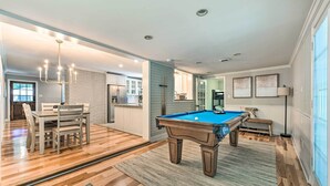 A spacious living room with a pool table, wooden flooring, a dining area, and a kitchen in the background.