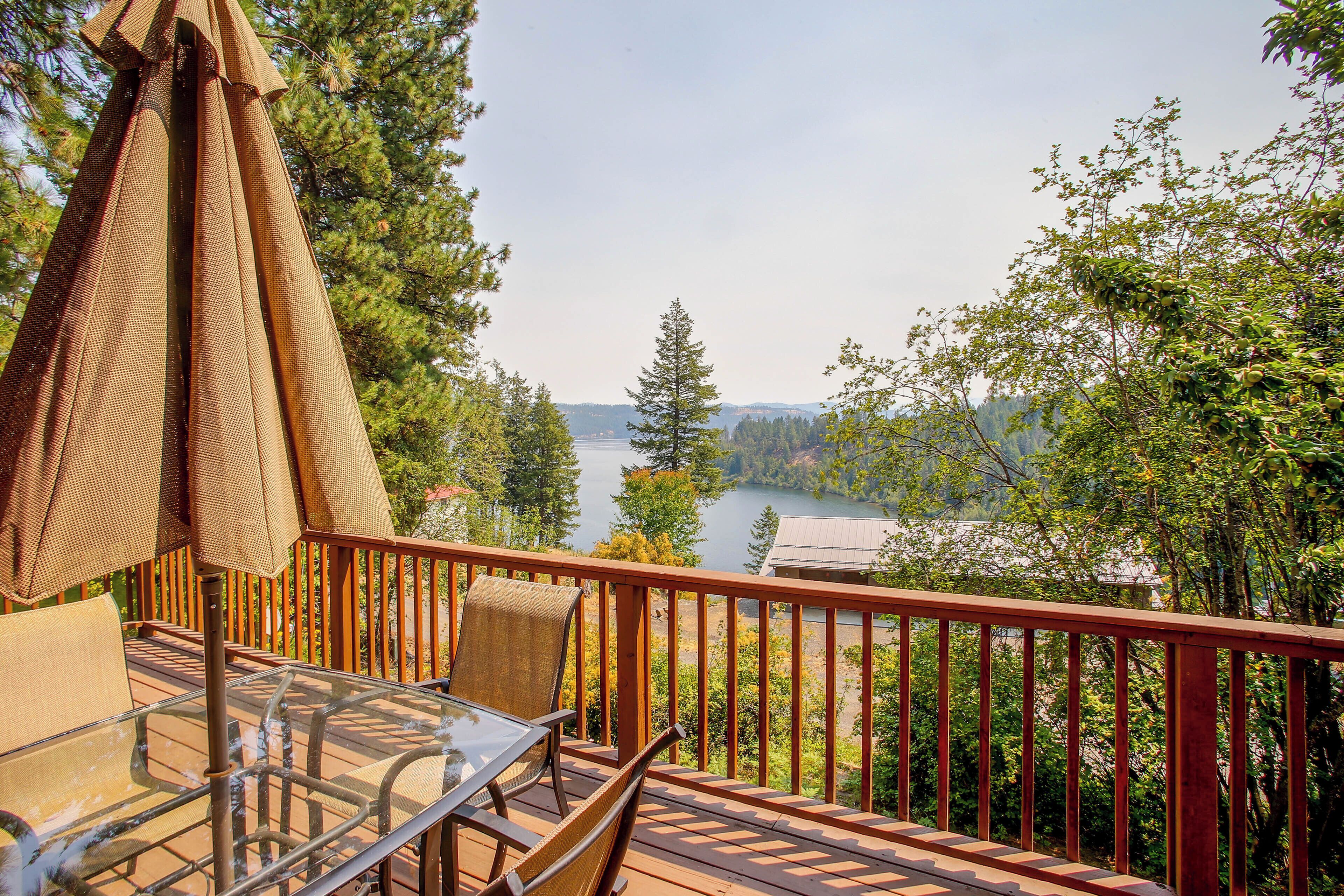 A deck with a glass table and chairs, overlooking a lake surrounded by trees.