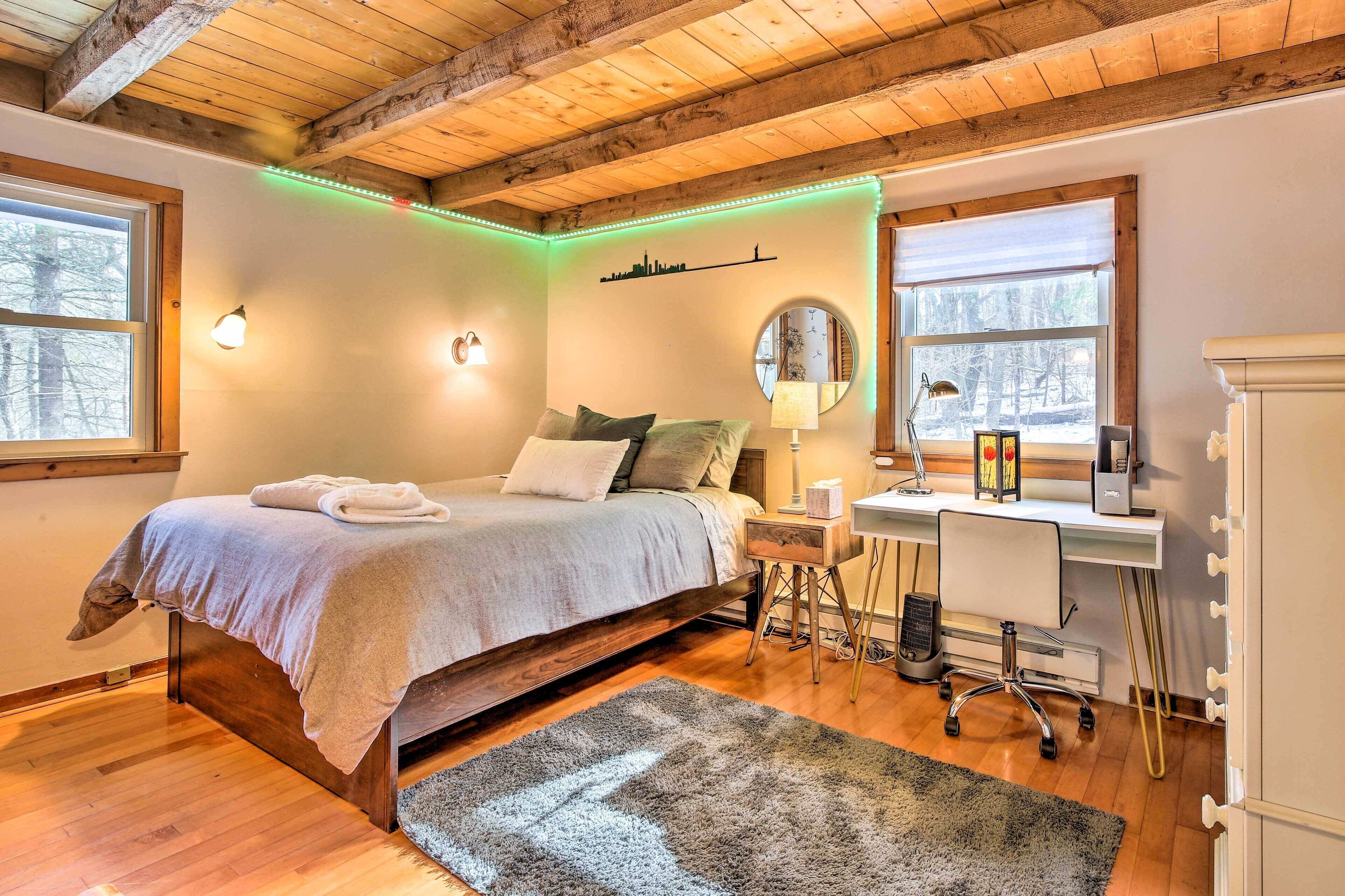 Ein Schlafzimmer mit Bett, Nachttisch, Lampe, Schreibtisch, Stuhl und Blick auf Bäume durch das Fenster.