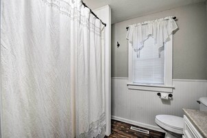 Un baño con inodoro blanco, ventana con persianas blancas y cortina de ducha.