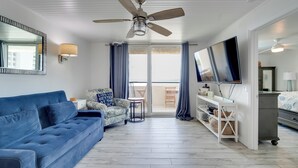 Un salon avec un canapé bleu, un fauteuil à motifs, un ventilateur de plafond, un téléviseur et une porte-fenêtre coulissante donnant sur un espace extérieur.