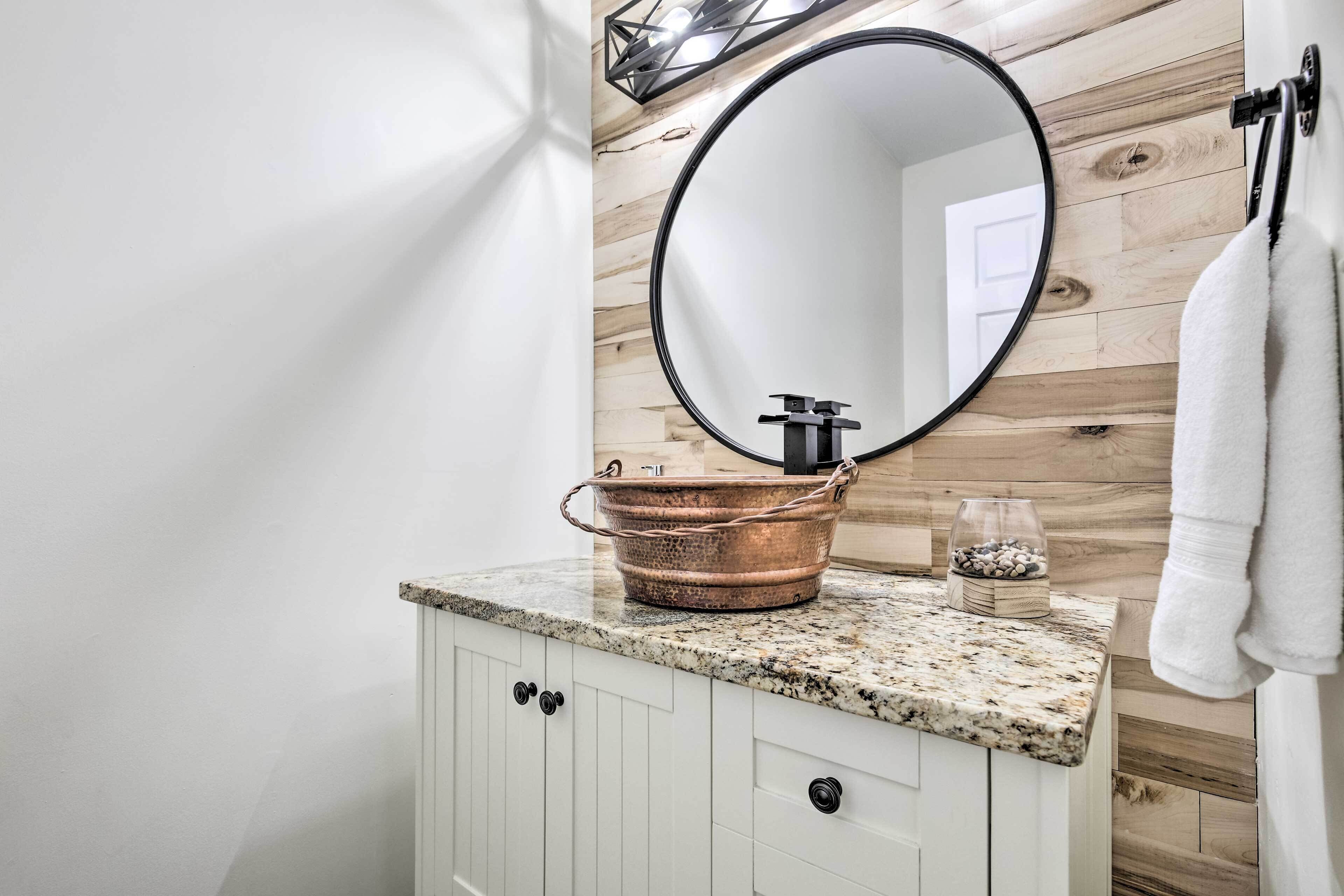 A bathroom with a wooden wall, a round mirror, a granite countertop, a copper basin, and a glass container.