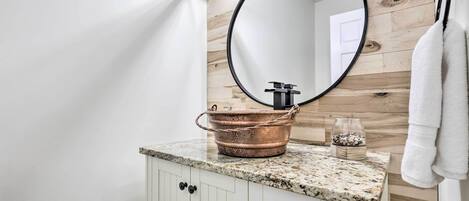 A bathroom with a wooden wall, a round mirror, a granite countertop, a copper basin, and a glass container.