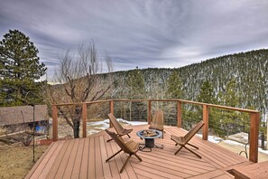 Una terraza de madera con mobiliario exterior y un hogar para fuego, rodeada por una barandilla de metal y con vistas a una zona boscosa.