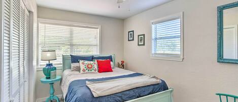A bedroom with a bed, a ceiling fan, two windows with blinds, a small table with a lamp, and a chair.