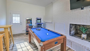 A pool table with a blue felt surface, a fireplace with a potted plant, and two arcade machines in the background. - Family-friendly New Bern Home w/ Backyard! (New Bern)