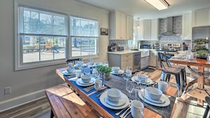 A modern kitchen with white cabinets, a wooden dining table set for six, and a large window overlooking a playground. - Family-friendly New Bern Home w/ Backyard! (New Bern)