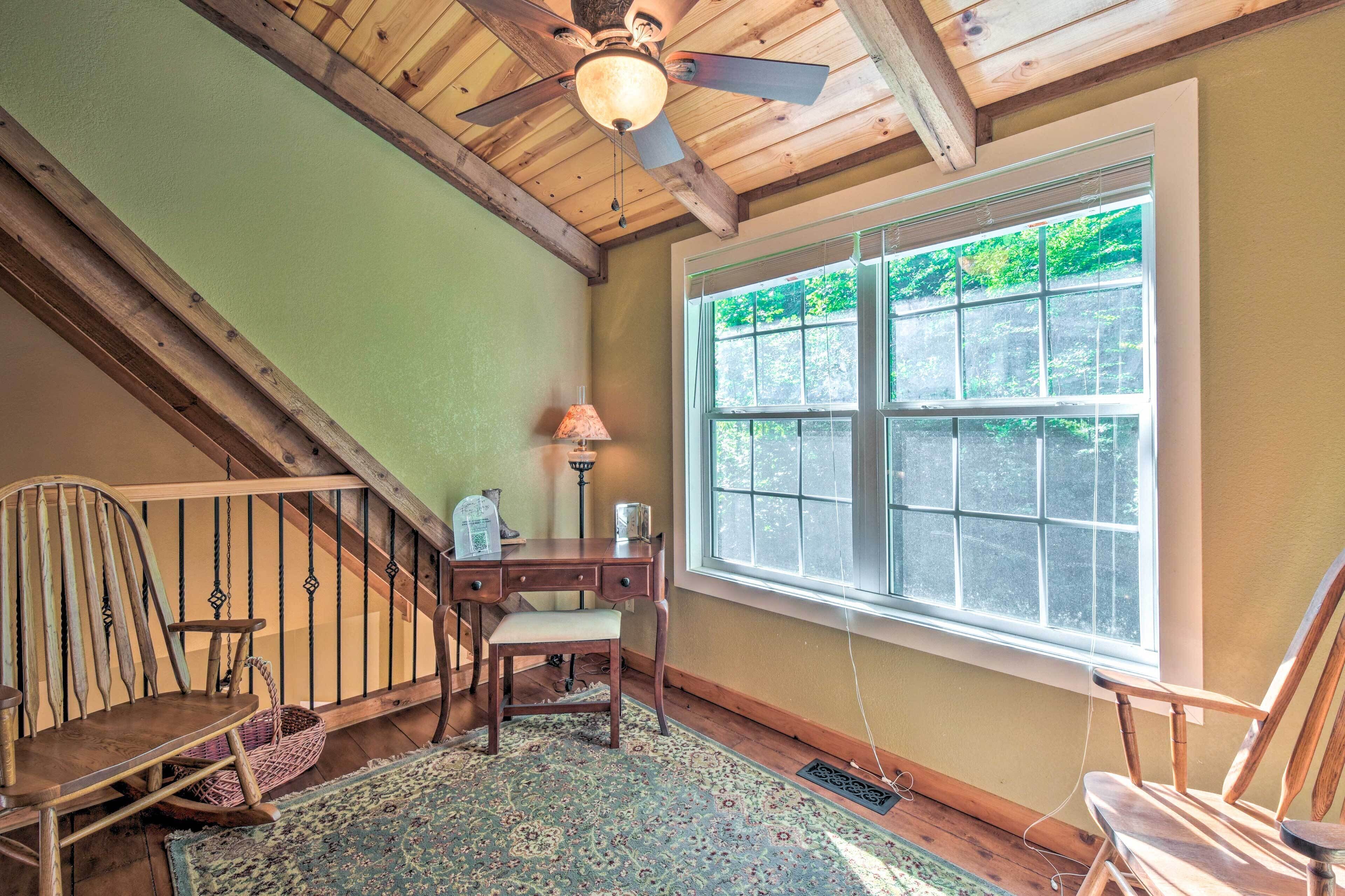 A room with a wooden ceiling, a large window with a view of greenery, a desk with a lamp, and a chair.