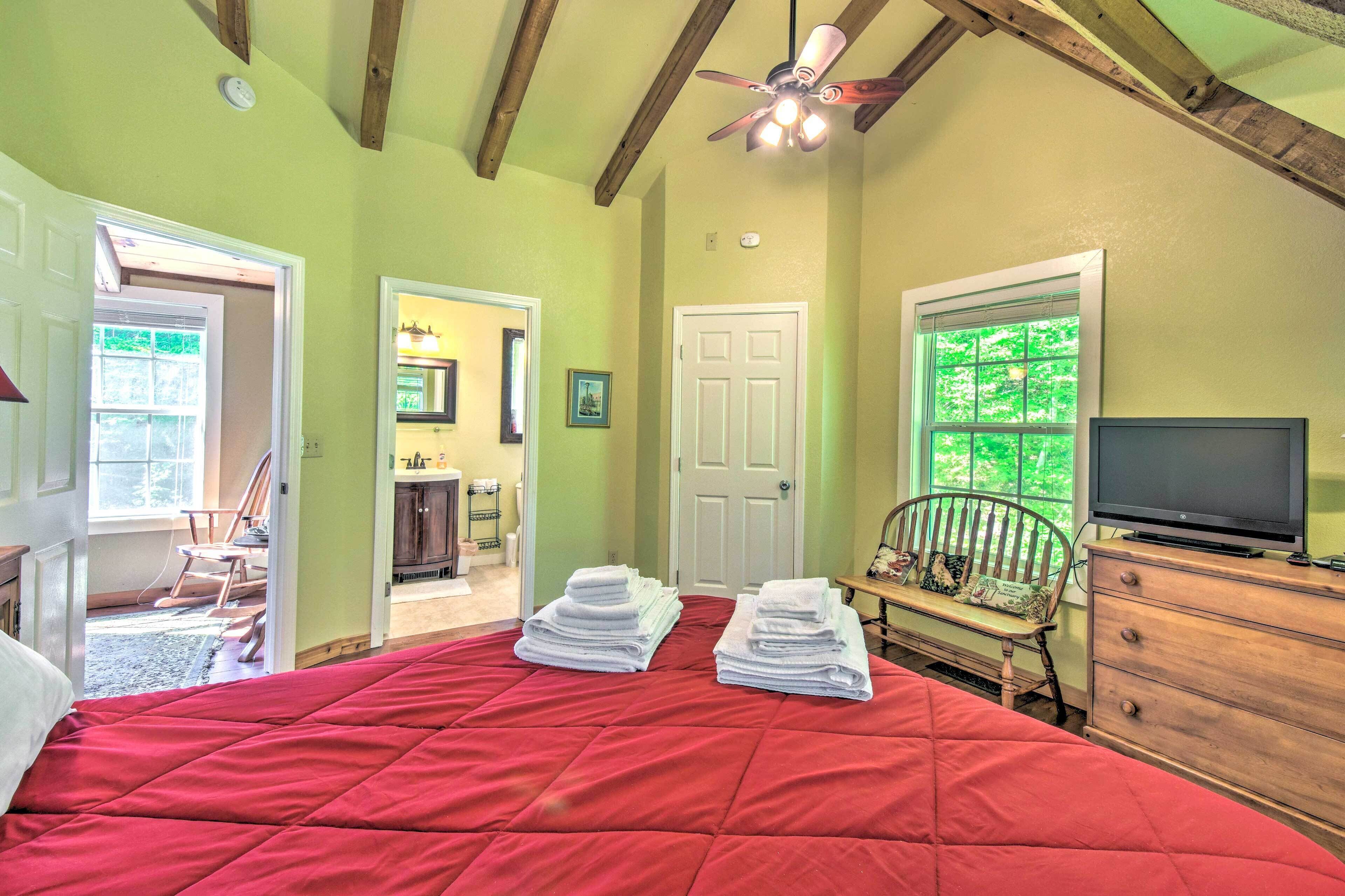 A bedroom with a red bedspread, a wooden dresser, a television, and a window with a view of trees.