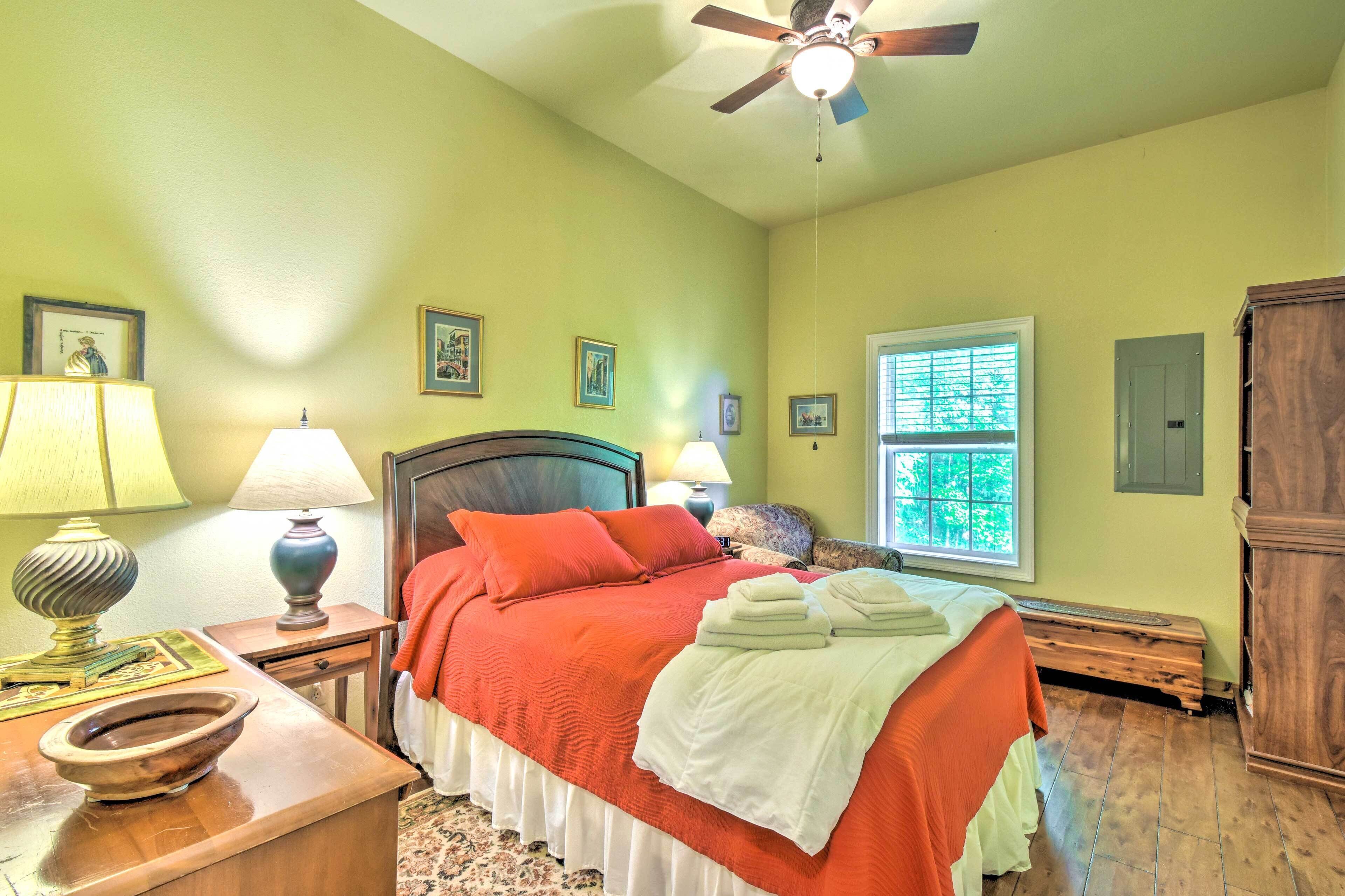 A bedroom with a bed, two bedside tables with lamps, a wooden nightstand, a window with blinds, and a ceiling fan.