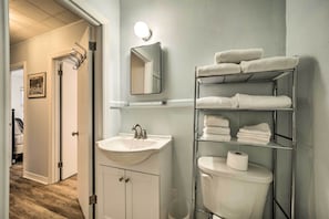 A bathroom with a white sink, mirror, and toilet. There is a towel rack with towels and a shelf with more towels.