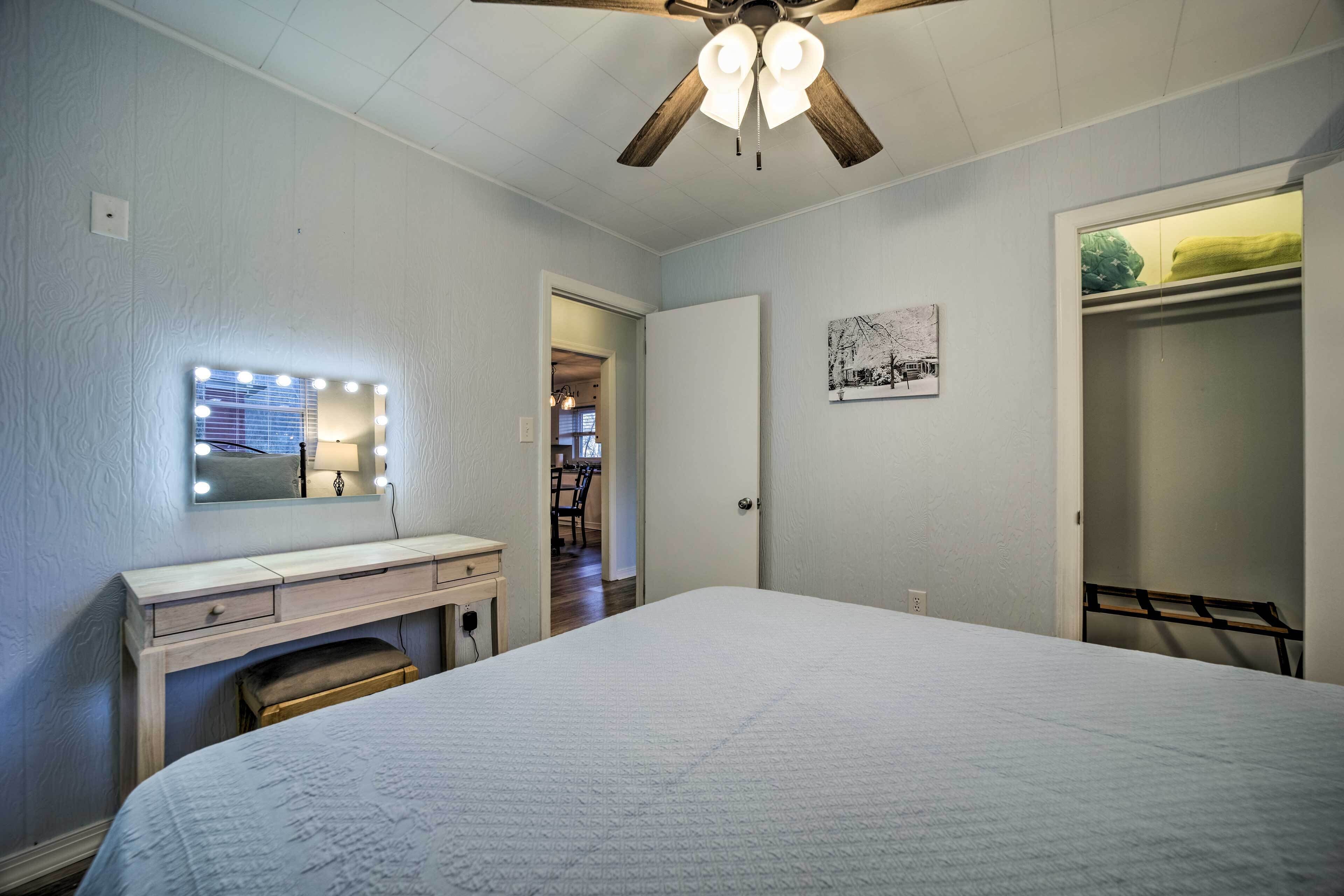 A bedroom with a bed, a vanity with a mirror and lights, a ceiling fan, and a door leading to another room.