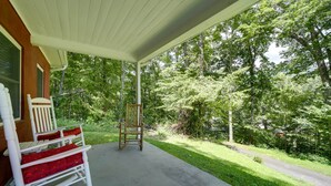 A covered porch with a rocking chair and a wooden chair, surrounded by lush greenery. - Maggie Valley Cottage Near Festival Grounds! (Maggie Valley)